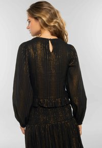 Blusa negra con un patrón de rayas doradas, mangas largas abullonadas, dobladillo con volantes y una pequeña abertura en forma de ojo de cerradura en la parte posterior. Tejido suave y texturizado.