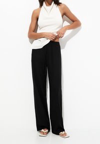 Top blanco sin mangas con cuello alto, combinado con pantalones anchos negros. Footwear presenta tiras blancas y un diseño de punta cuadrada.
