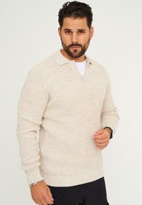Beige gestrickter Pullover mit Kragen, gerippten Bündchen und Saum, der ein strukturiertes Muster und eine lockere Passform aufweist.