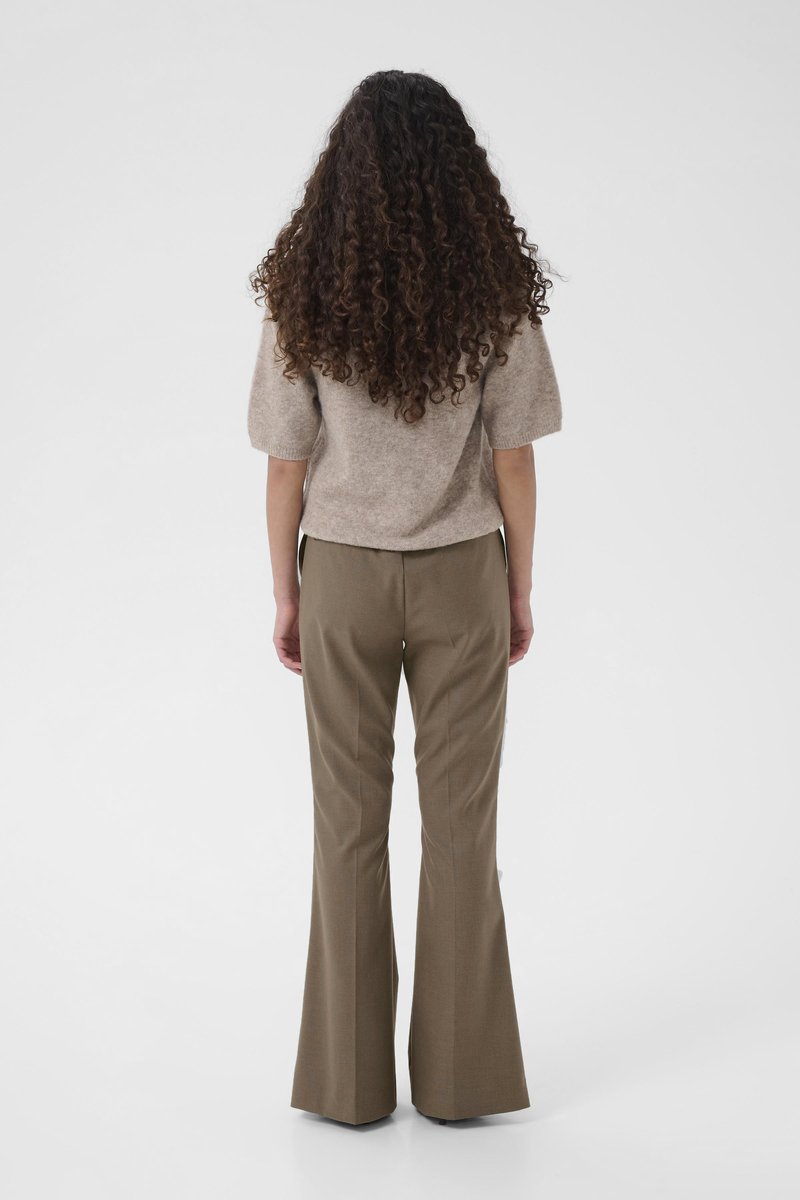Pull en mélange de laine beige à manches courtes, associé à un pantalon taille haute vert olive avec des ourlets évasés. Cheveux bouclés et longs.