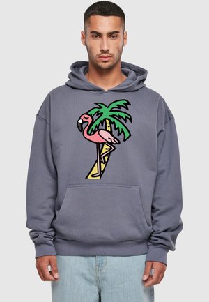 Man in een blauwgrijze hoodie met een kleurrafische afbeelding van een roze flamingo die op één been staat voor een groene palmboom.