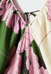 Gros plan sur un tissu fleuri avec des fleurs roses, des feuilles vertes et une partie crème, présentant un col en V et un détail de cordon noir noué.