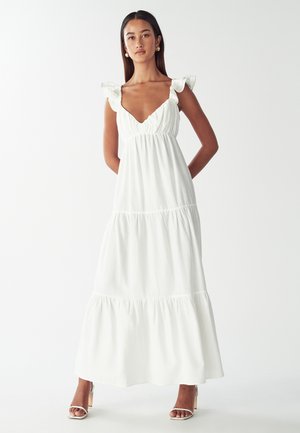Weißes Maxikleid mit Rüschenträgern, tiefem V-Ausschnitt, geraffter Taille, gestuftem Rock und einem leichten, strukturierten Stoff. Kombiniert mit Riemchen-Heels.