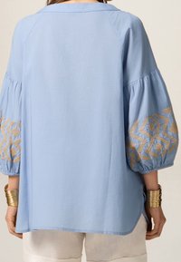 Jasnoniebieska bluzka z balonowymi rękawami z złotymi haftowanymi wzorami. Miękka tkanina, luźny krój i zakrzywiony fason.