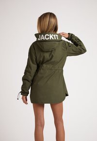 Kvinde i olivengrøn hættejakke med "JACK1T" tekst på hætten, vist fra ryggen, mens hun retter hætten til med den ene hånd.