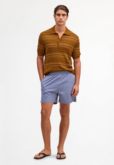 Young man standing with hands in pockets, wearing a brown patterned short-sleeve polo shirt, light blue shorts, and black flip-flops.