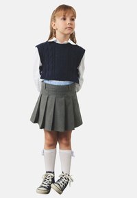 Barn iført en navy strikket vest over en hvid skjorte, grå plisseret nederdel, knæhøje hvide strømper og navy sneakers med hvide såler.