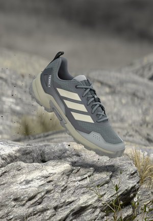 Γκρι και μπεζ παπούτσι Adidas Terrex για τρέξιμο σε μονοπάτια στον αέρα πάνω από πετρώδες έδαφος, που αναδεικνύει τη σκληρή σόλα και το δίχτυ στο πάνω μέρος.