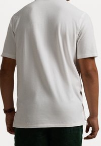 Bílý krátký rukáv tričko s texturovaným povrchem, zakulaceným lemem a bočními rozparky. Materiál je lehký a prodyšný.