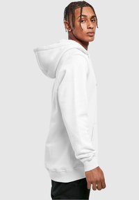 Hvid hoodie med en rummelig hætte, frontlomme og ribbede manchetter. Glat tekstur, løs pasform, minimalistisk design. Modellen vist i profil.