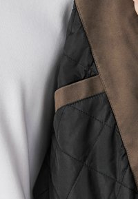 Gros plan d'une veste matelassée noire avec col en daim marron et sangle sur un fond de tissu blanc.