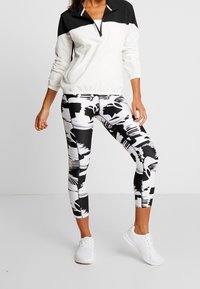 Chaqueta de cremallera en blanco y negro con un cuello alto, combinada con leggins estampados que presentan formas abstractas y zapatillas blancas.