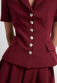 Giacca da donna tailor in bordeaux a maniche corte con sei bottoni decorativi in perla e tonalità dorata, tasche a soffietto e gonna plissettata coordinata.