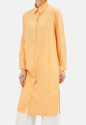 Frau trägt ein langärmliges, pfirsichfarbenes Hemdkleid mit Knöpfen und Seitenschlitzen über weißen Hosen vor einem schlichten Hintergrund.