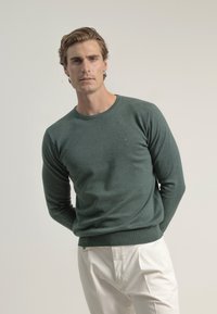Suéter verde hecho de tejido de punto suave, con un escote redondo, mangas largas y un logo sutil en el pecho. Combinado con pantalones blancos.
