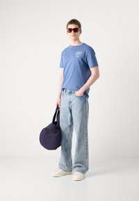 Modra bombažna majica s logotipom, svetlo modri izbližani široki jeans, beli superge in okrogla mornarsko modra torba z zlatim detajlom. Vključene so sončna očala.