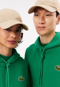 Sudaderas verdes de algodón con cordones y un pequeño logo bordado. Gorras beige completan el look. El diseño es sencillo y casual.