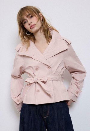 Ženska nosi svetlo roza plašč s pasom in velikim ovratnikom ter temno modre hlače, pózira z rokami v žepih pred enobarvnim ozadjem.