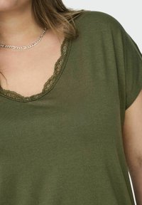 Olivovo zelené tričko s výstrihom do tvaru lopatky, s dekoratívnym čipkovým lemovaním. Mäkkučký, ľahký materiál a krátke rukávy.