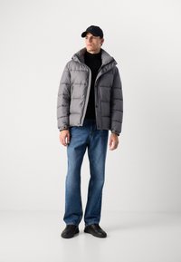 Gray puffer jacket with a high collar, button closure, and patch pockets, paired with blue jeans and black sneakers. Black cap completes the look.