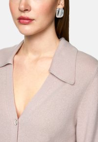 Hellrosa Strickjacke mit V-Ausschnitt, weicher Textur und einem Kragen mit silbernen Akzenten. Das Model trägt große silberne Ohrringe.