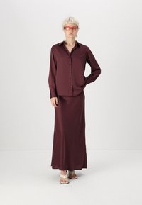 Satinová burgundy košeľa a dlhá sukňa, ktorá sa skladá z košele s golierom a gombíkmi a plochých sandálov, doplnená červenými slnečnými okuliarmi.