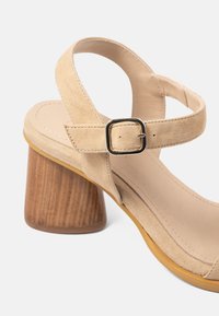 Sandală din suede bej cu toc din lemn, curea de gleznă ajustabilă și cataramă rectangulară aurie pe un fundal alb.