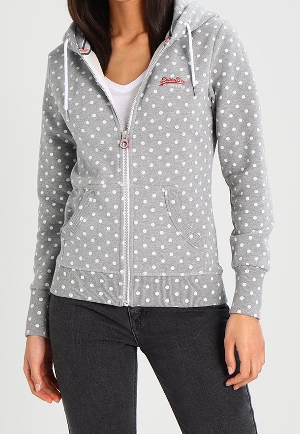 Femme portant un sweat à capuche gris zippé avec des pois blancs, des cordons blancs et un logo rouge sur la poitrine, par-dessus un t-shirt blanc.