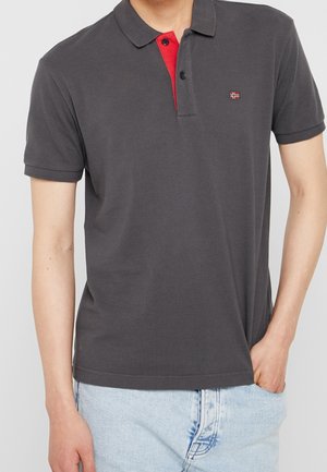 Mężczyzna ubrany w ciemnoszarą koszulkę polo z krótkim rękawem z czerwonym detalem przy guzikach i małym logo flagi, jasnoniebieskie dżinsy, z jedną ręką w kieszeni.