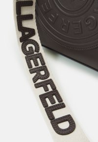 Bruine leren handtas met een gestructureerd cirkelpatroon, voorzien van een beige band met de geperste letters "LAGERFELD" in donkerbruine kleur.