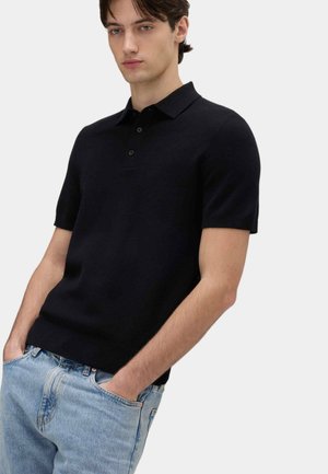 Ung mand iført en sort kortærmet polotrøje og lyseblå jeans med hænderne i lommerne, stående mod en ensfarvet baggrund.