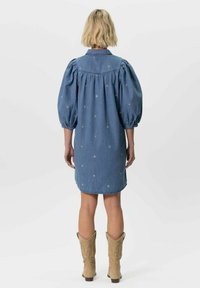 Robe en denim avec des manches bouffantes, un empiècement dans le dos et un motif d'étoiles brodé. Mi-longue avec une légère forme en A. Portée avec des bottes de cowboy beiges.