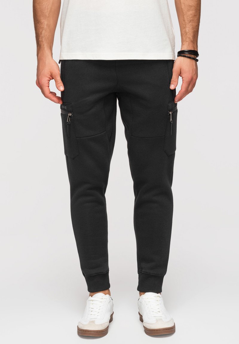 Homme portant un jogger cargo noir slim avec des poches zippées et des baskets blanches, debout devant un fond uni.