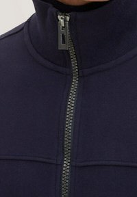 Giacca in pile blu navy con colletto alto, zip a lunghezza completa con tirazipp in metallo e cuciture piatte sul pannello anteriore.
