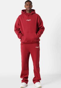 Conjunto de sudadera y pantalones deportivos en color burdeos, fabricado con tejido suave, que incluye un bolsillo frontal, capucha con cordón y acentos en blanco del logotipo en el pecho y el muslo.