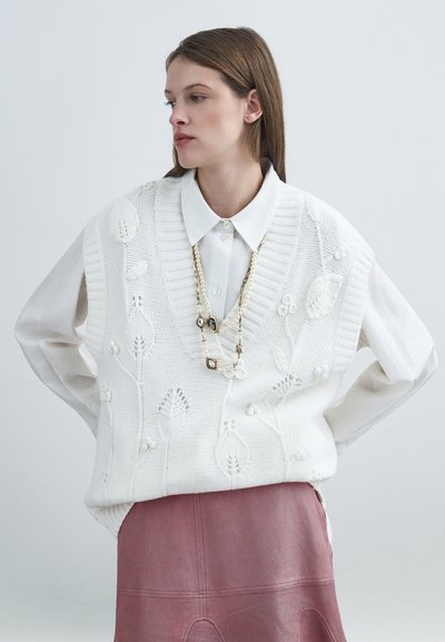 Veste blanche en tricot avec broderie florale, portée par-dessus une chemise légère. Détails notables : col en V et délicat collier de perles. Jupe en cuir rose.