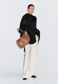 Žena stojící v černém oversized kabátu, bílých širokých kalhotách, černých botách a nese velkou hnědou koženkovou tašku.