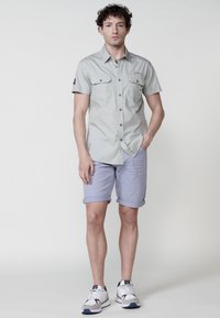 Maglietta grigia chiara a maniche corte con due tasche frontali, abbinata a pantaloni corti azzurri arrotolati e scarpe da ginnastica bianche. Design e tessuto semplici.