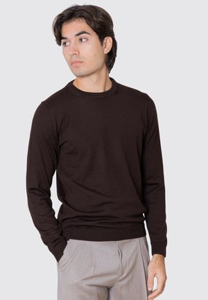 Giovane uomo con capelli scuri che indossa un maglione nero a maniche lunghe e pantaloni grigio chiaro, guarda verso sinistra con una mano in tasca.