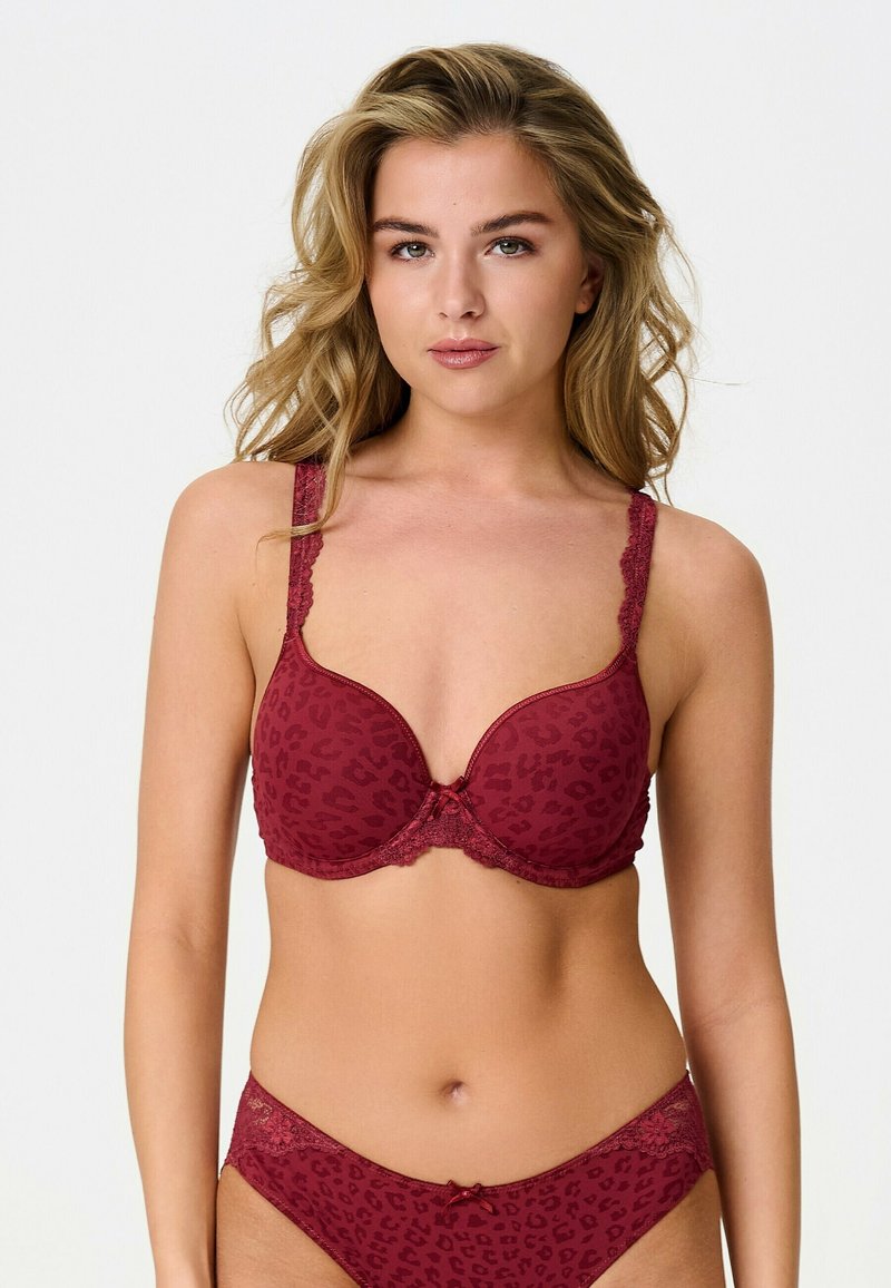Femme aux cheveux blonds ondulés portant un soutien-gorge et une culotte assortis imprimés léopard rouge foncé avec des détails en dentelle, posant devant un fond clair uni.