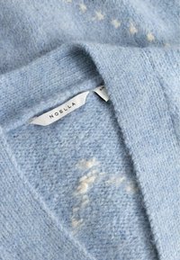 Vaaleansininen neulottu pusero pehmeällä tekstuurilla, pienillä valkoisilla tähtikuviolla ja näkyvällä vaatemerkinnällä, jossa lukee "NOELLA".