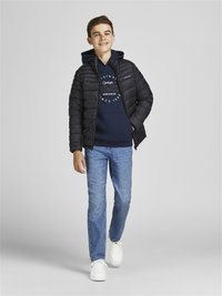 Giacca a vento nera sopra un hoodie blu navy con testo grafico, abbinata a jeans blu e sneaker bianche. Modella che cammina con uno sfondo neutro.