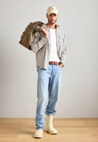 Casquette beige, chemise à carreaux bleu et jaune, t-shirt blanc, jean bleu clair, ceinture marron, et bottes imperméables beiges. Portant un sac marron.