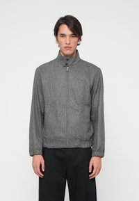 Grå zip-up jakke med høj krave, struktureret stof og subtilt mønster. Har elastiske manchetter og et broderet logo på brystet.
