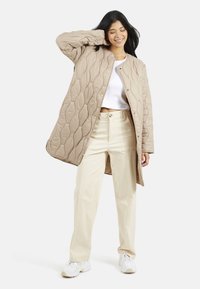 Cappotto lungo beige trapuntato con un motivo testurizzato, bottoni a scatto e un collo rotondo, abbinato a pantaloni chiari e sneaker bianche.