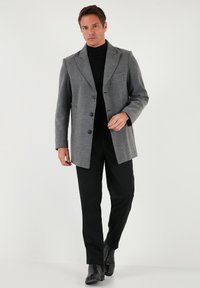 Sivi plašč iz volnene mešanice z izrezanimi ovratniki, enodelno zapenjanje na gumbe in sprednjim žepom. Nošen preko črnega polo-obleke in črnih hlač.