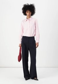 Camicia button-up rosa chiaro con strisce verticali, abbinata a pantaloni larghi blu navy. Scarpe nere a punta e una borsa rossa completano il look.
