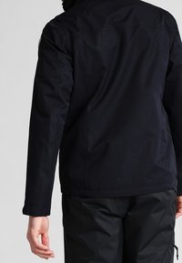 Námořnická modrá nepromokavá bunda s vysokým límcem, přiléhavým střihem a mírnou texturou. Obsahuje logo na levém rameni a bezešvé provedení zad.