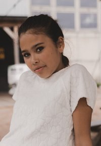 Witte textuur top met bloemenpatroon, korte mouwen. Glad stof, relaxte pasvorm. Achtergrond heeft een zachte focus en buitenelementen.