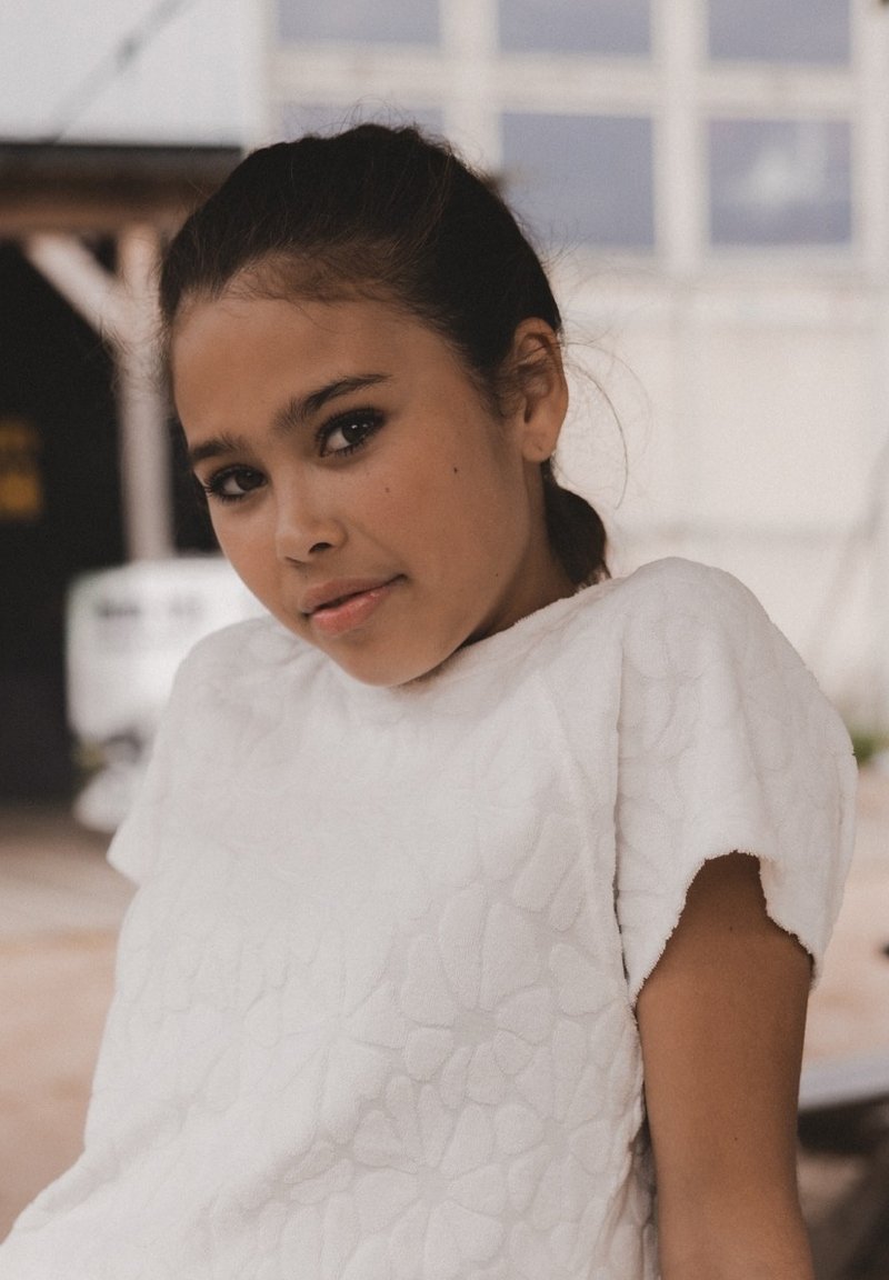 Witte textuur top met bloemenpatroon, korte mouwen. Glad stof, relaxte pasvorm. Achtergrond heeft een zachte focus en buitenelementen.